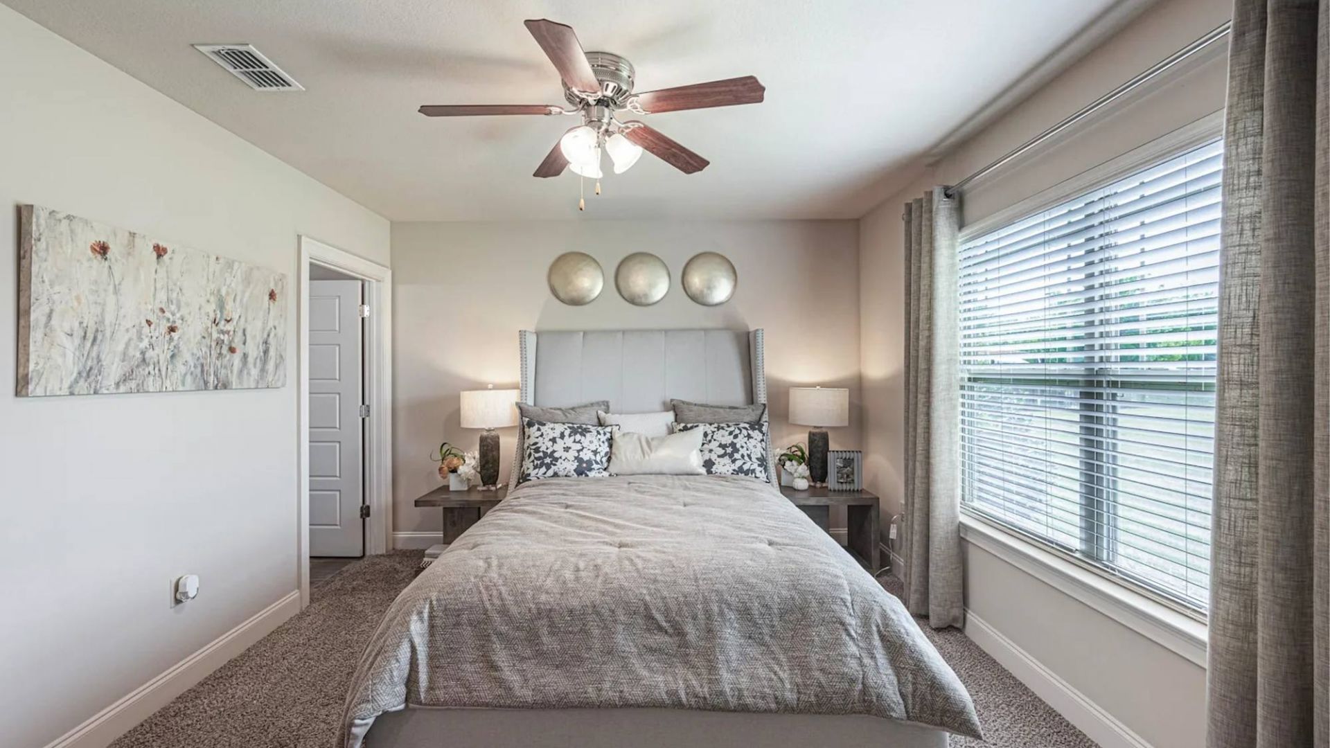 The Grove at Stone Park apartment homes with Modern bedroom with gray bedding, large window, ceiling fan, and wall art above the bed.