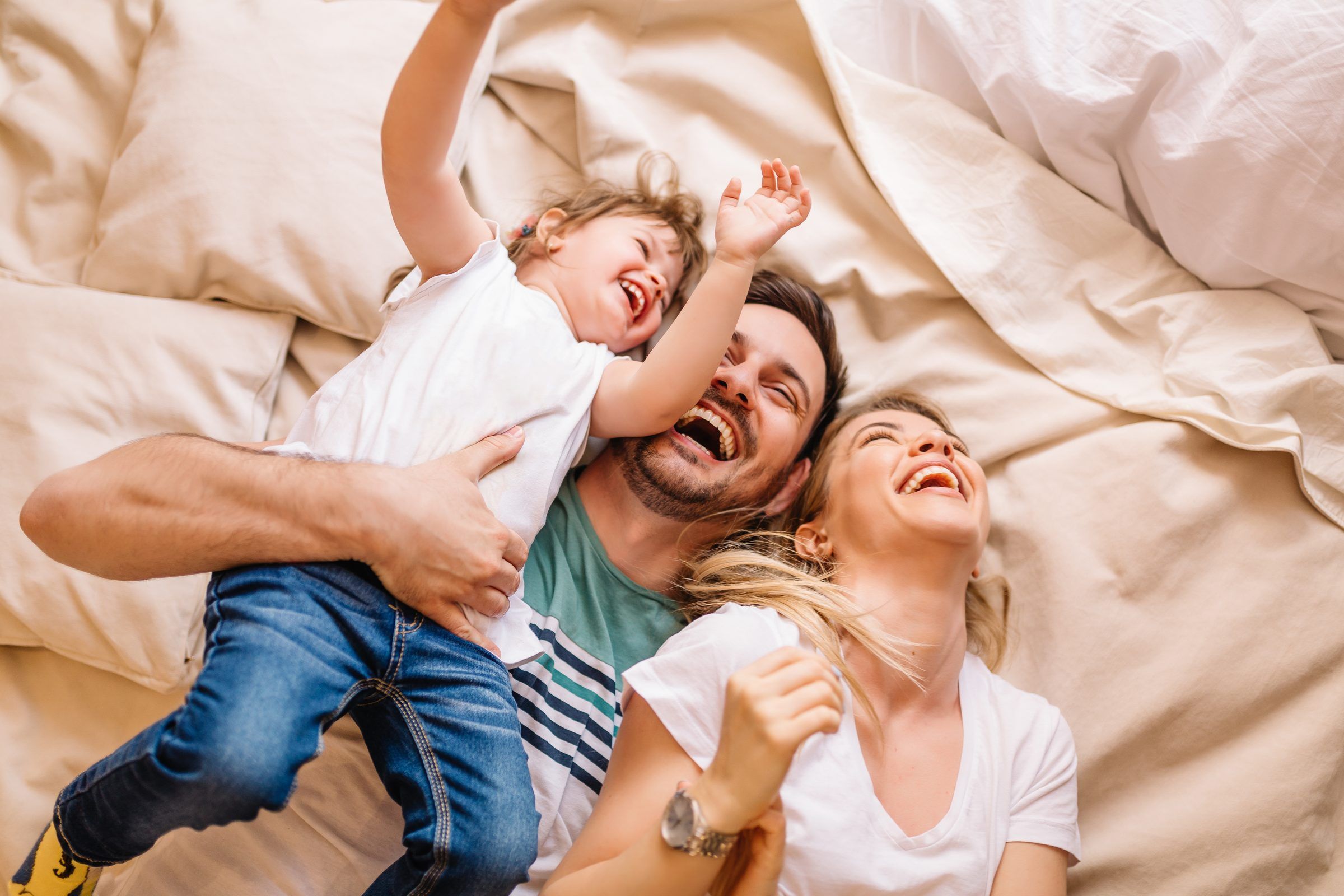 The Grove at Stone Park apartment homes with A joyful family of three laughs together while lying on a bed with cream-colored sheets.