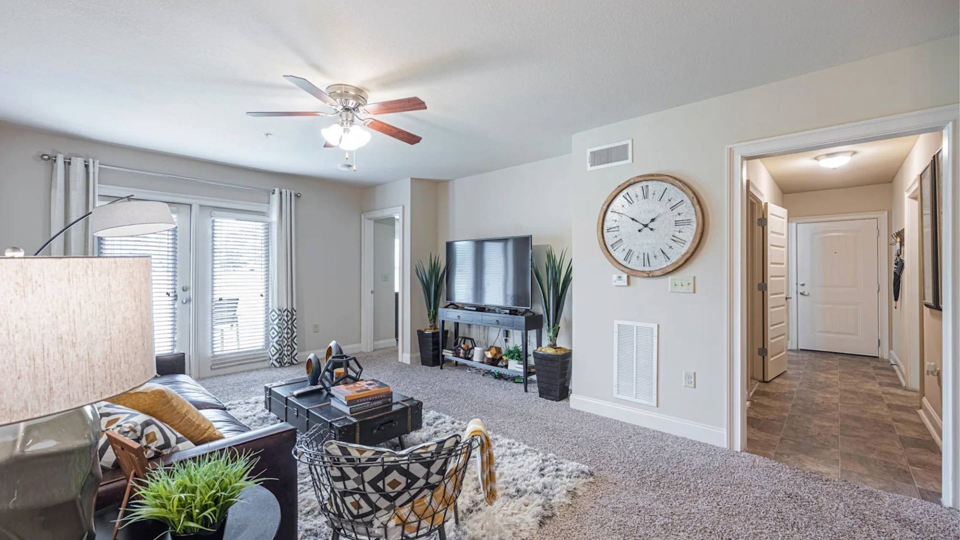 The Grove at Stone Park apartment homes with Bright living room with modern decor, TV, large clock, and glass doors leading to a balcony.