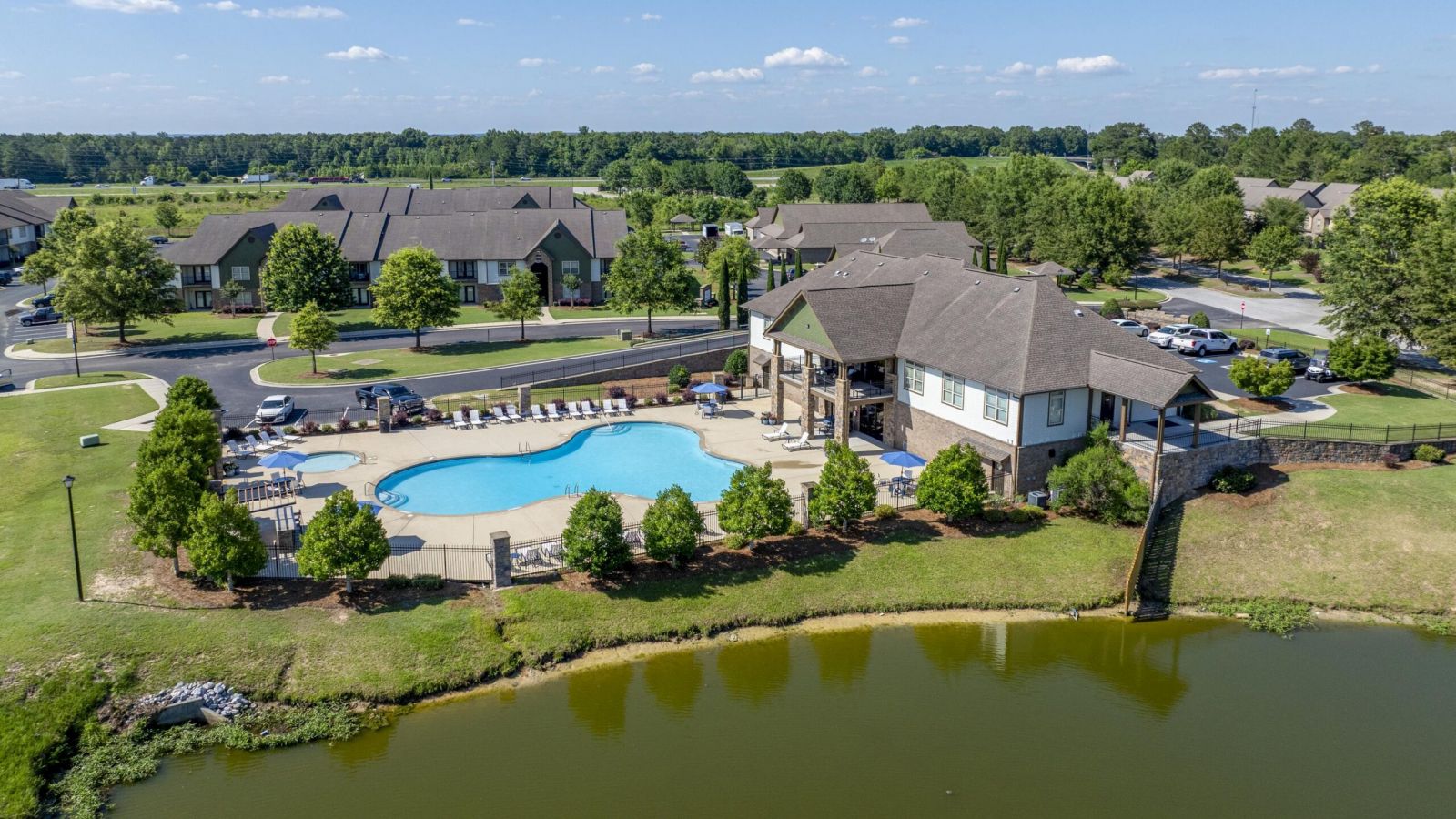 The Grove at Stone Park apartment homes with Aerial view of a community pool, clubhouse, and pond surrounded by houses, trees, and parked cars on a sunny day.