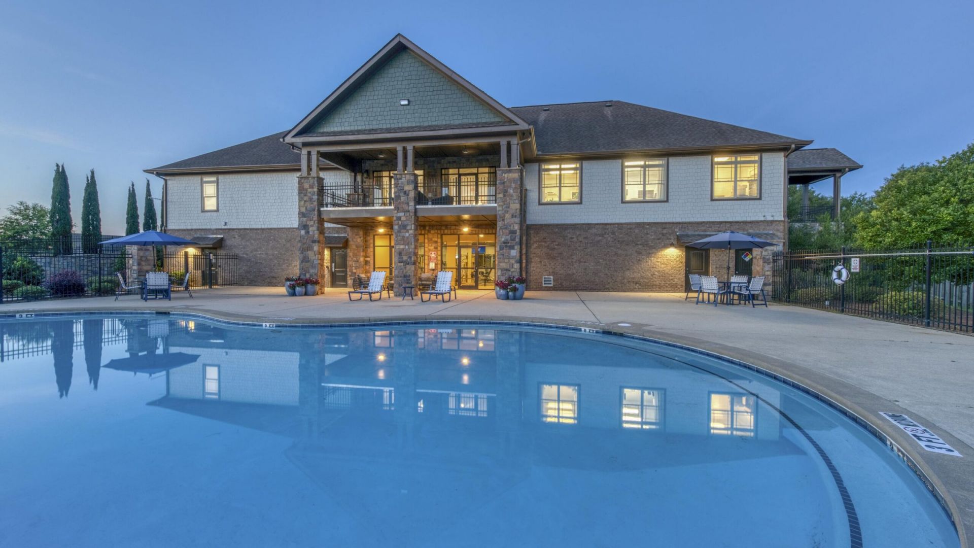 The Grove at Stone Park apartment homes with Large house with lit windows, patio seating, and a clear blue swimming pool in the foreground at dusk.