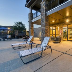 The Grove at Stone Park apartment homes with Patio with white lounge chairs, potted flowers, and stone columns near glass doors at dusk; poolside area visible on the left.
