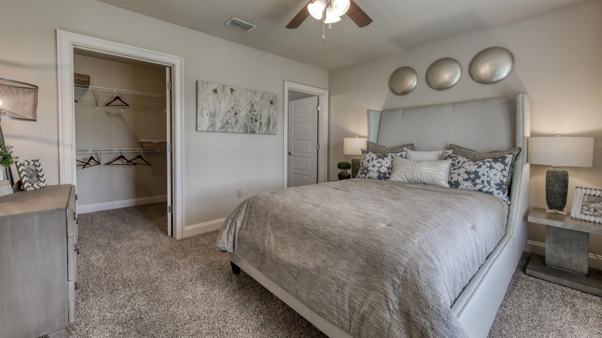 The Grove at Stone Park apartment homes with A modern bedroom with a gray bed, patterned pillows, nightstands, lamps, and an open walk-in closet with shelves and hangers.