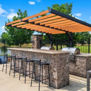 The Grove at Stone Park apartment homes with Outdoor brick grill area with bar seating, a pergola, and a scenic view of trees and a pond in the background.