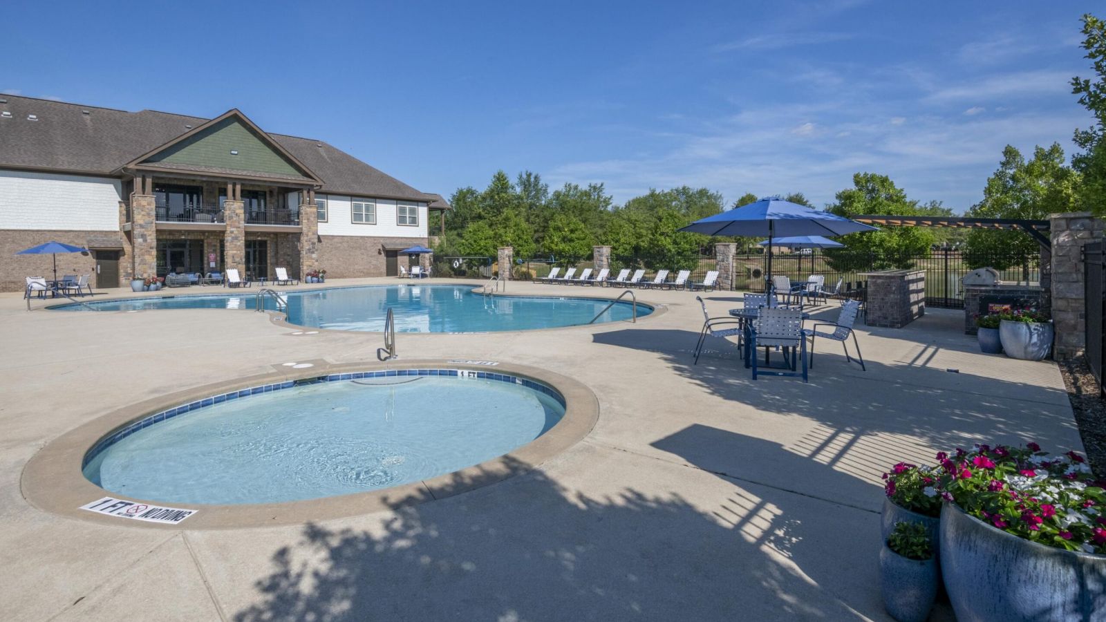 The Grove at Stone Park apartment homes with Outdoor pool area with lounge chairs, tables with umbrellas, a smaller pool, and a building in the background under a clear blue sky.