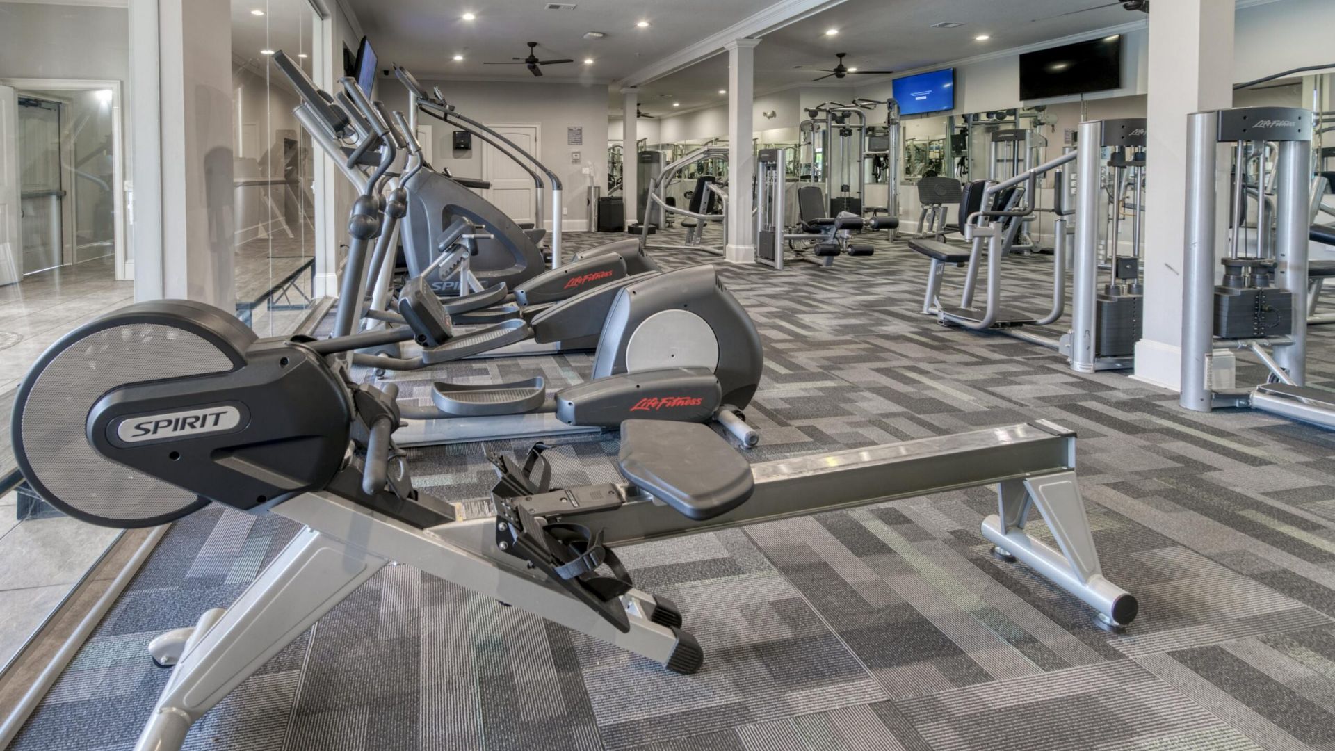 The Grove at Stone Park apartment homes with Modern gym with cardio machines, weight equipment, mirrors, ceiling fans, and gray patterned carpet. The room is brightly lit and spacious.